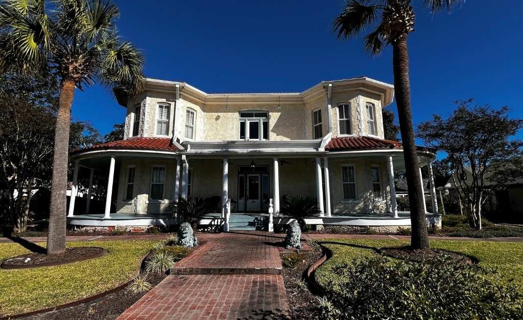Wide exterior view of The Caldwell House Bed and Breakfast in Abbeville, Louisiana, showing the symmetrical facade, palm trees, and brick walkway.