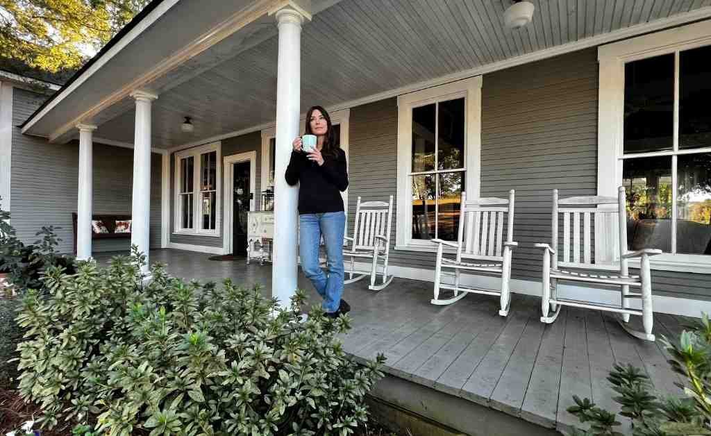 Karen LeBlanc standing on the front porch of the Dover House in Florien, Louisiana, holding a mug beside white rocking chairs